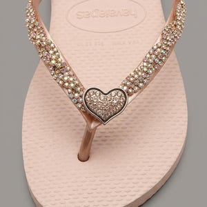 May include: A light pink flip-flop sandal featuring a braided strap adorned with iridescent crystals. A central heart-shaped charm, also covered in small crystals, sits at the junction of the straps. The footbed displays the Havaianas brand logo.