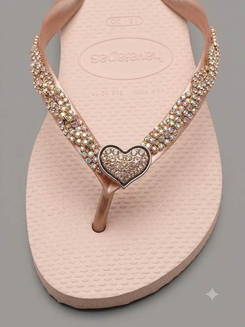 May include: A light pink flip-flop sandal featuring a braided strap adorned with iridescent crystals. A central heart-shaped charm, also covered in small crystals, sits at the junction of the straps. The footbed displays the Havaianas brand logo.