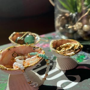 Pode incluir: Dois pequenos recipientes para joias em forma de concha branca com bordas pintadas de dourado. Um recipiente contém uma pulseira de contas brancas e verde-claras, enquanto o outro tem brincos de argola dourados. Outra concha contém contas verdes.