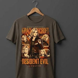 May include: A dark gray t-shirt featuring a graphic design with the text "GRACEHCROFT" and "RESIDENT EVIL REQUIEM". The design includes multiple images of a woman with blonde hair. The shirt is hanging on a wooden hanger.