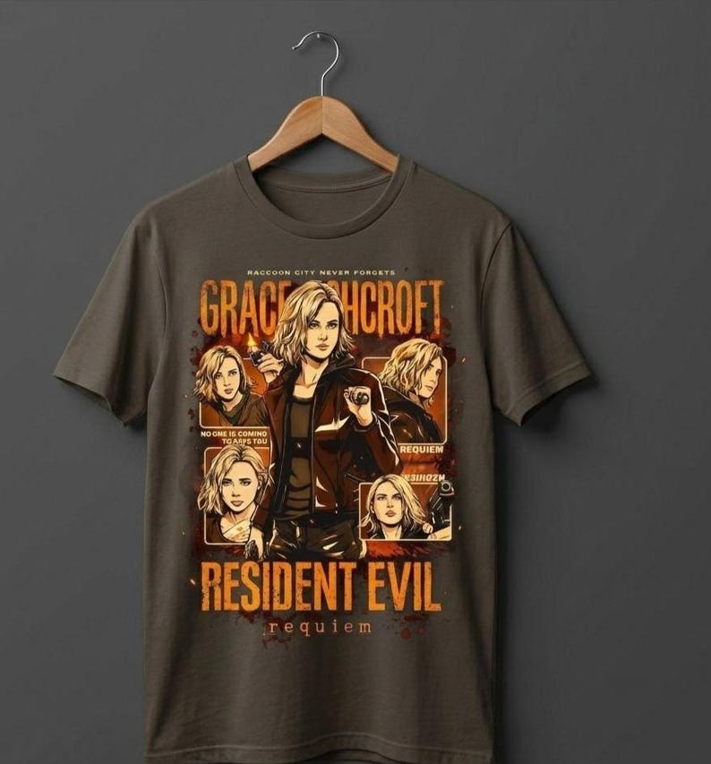 May include: A dark gray t-shirt featuring a graphic design with the text "GRACEHCROFT" and "RESIDENT EVIL REQUIEM". The design includes multiple images of a woman with blonde hair. The shirt is hanging on a wooden hanger.