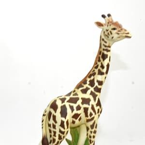 Op de afbeelding: Een keramiek giraffenbeeldje met bruine en witte vlekken dat op een groene en bruine basis staat. De giraffe kijkt naar rechts en heeft een lange nek.