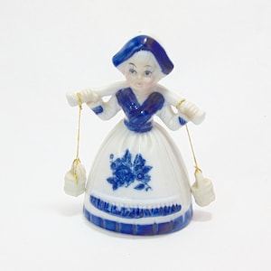 Puede incluir: Figura de cerámica de una mujer con vestimenta tradicional holandesa. El vestido blanco presenta detalles florales y rayas azules. La figura lleva dos cubos blancos suspendidos por cuerdas doradas. El sombrero y el corpiño son azules.