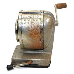 Puede incluir: Sacapuntas vintage de metal plateado y gris con manivela y montaje de vacío. El sacapuntas tiene las palabras "BOSTON VACUUM MOUNT" impresas en el lateral.