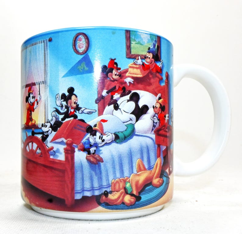 Puede incluir: Una taza de cer&aacute;mica blanca con una ilustraci&oacute;n colorida de Mickey Mouse y sus amigos en un dormitorio. Mickey est&aacute; dormido en la cama, mientras los otros personajes juegan a su alrededor. La taza tiene un borde y un asa azules.