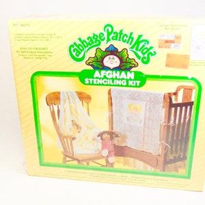 Puede incluir: Una caja de kit de esténcil para afganistán de Cabbage Patch Kids con fondo amarillo y verde. La caja muestra una mecedora, una cuna y una muñeca. El texto en la caja dice "Cabbage Patch Kids Afghan Stenciling Kit".
