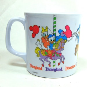 May include: A white ceramic mug with a colorful illustration of Donald Duck, Daisy Duck, and a horse on a carousel. The mug has the text "Disneyland" and "©DISNEY" printed on it.