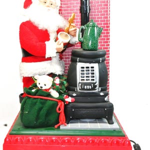 Può includere: Decorazione natalizia animata con Babbo Natale in abito e cappello rossi, che tiene in mano una tazza e un biscotto. Una teiera verde è posta su una stufa nera accanto a un camino in mattoni. Un sacco verde con un orsacchiotto e bastoncini di zucchero è alla base.