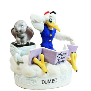 May include: A ceramic figurine featuring a gray elephant sitting in a light blue bundle next to a white stork wearing a blue vest and red cap. The stork holds a purple map labeled Map of Circus Route. The base is white with the text DUMBO.