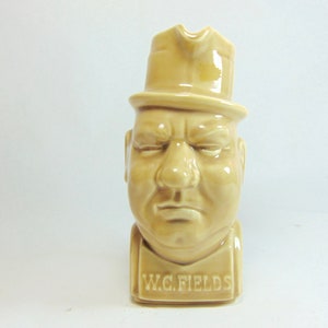 May include: A ceramic pitcher shaped like the head of a man wearing a hat. The pitcher is beige and has the words "W.C. FIELDS" written on the front.
