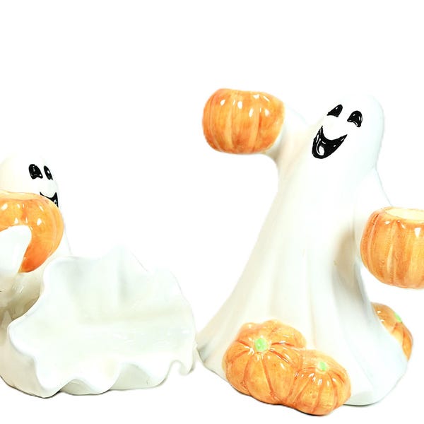 May include: Two white ceramic ghost figurines with black painted facial features. One ghost holds a single orange pumpkin, while the other stands with arms raised holding an orange pumpkin and has two additional orange pumpkins at its base.