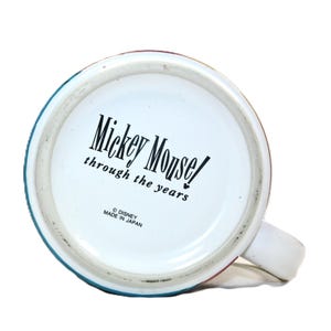Puede incluir: Taza de cer&aacute;mica blanca con un borde azul, rojo y verde. La parte inferior de la taza tiene el texto "Mickey Mouse! through the years" y "&copy; DISNEY MADE IN JAPAN".