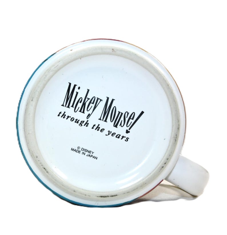 Puede incluir: Taza de cer&aacute;mica blanca con un borde azul, rojo y verde. La parte inferior de la taza tiene el texto "Mickey Mouse! through the years" y "&copy; DISNEY MADE IN JAPAN".
