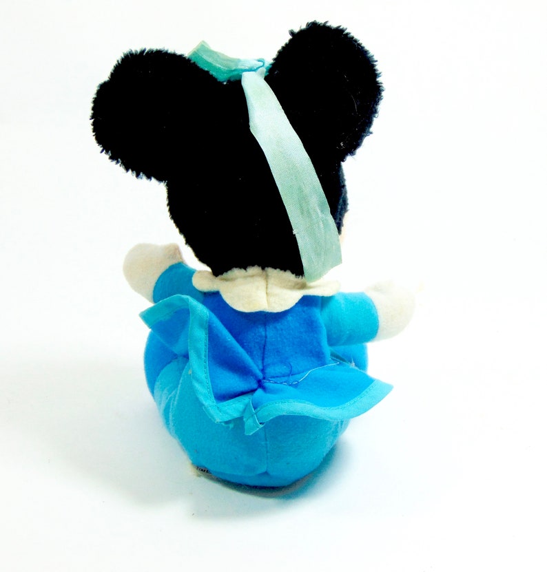 May include: A plush toy of a cartoon mouse wearing a blue dress with a white collar and a blue bow. The mouse has black ears and a black face.