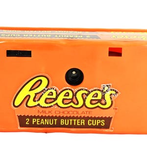 May include: A rectangular orange disposable camera featuring the Reese's Milk Chocolate 2 Peanut Butter Cups logo on the front. The camera has a central black lens, a black film advance wheel on the top left, and a red film counter window on the top right.