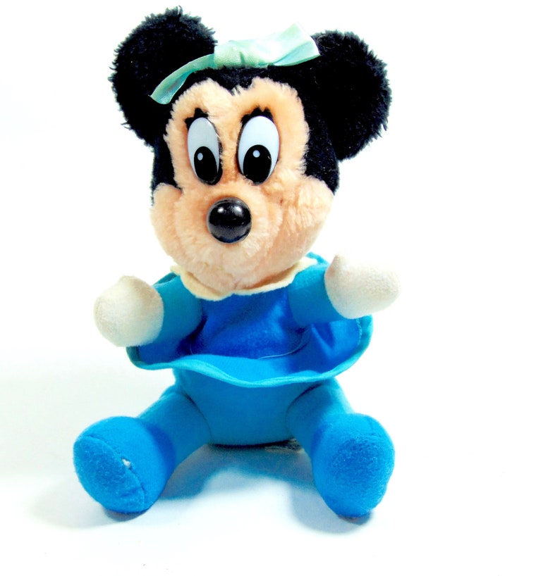 May include: A plush toy of Minnie Mouse wearing a blue dress and a green bow. She has black ears and a brown nose.
