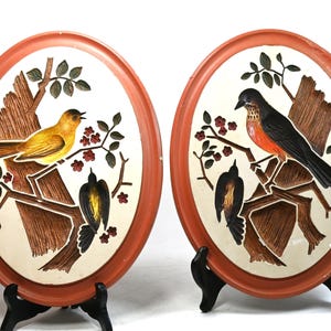 May include: Two oval wall plaques with a bird theme. Each plaque features a bird perched on a branch with leaves and flowers. The birds are yellow and black, and orange and black. The plaques have a brown border and are displayed on black stands.