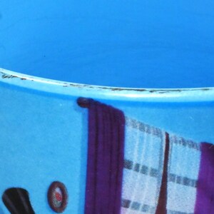 Puede incluir: Una taza de cer&aacute;mica azul con asa blanca. La taza tiene un dise&ntilde;o colorido con un reloj, cortinas y otros detalles.