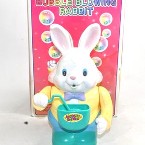 May include: A white rabbit toy with a yellow shirt, blue pants, and a green bucket. The toy blows bubbles and plays music. The box says "Musical Bubble Blowing Rabbit".
