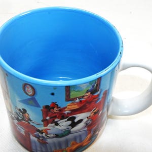 Puede incluir: Una taza de cer&aacute;mica blanca con interior azul y una imagen colorida de Mickey Mouse y sus amigos. La imagen muestra a Mickey Mouse y sus amigos en un dormitorio.