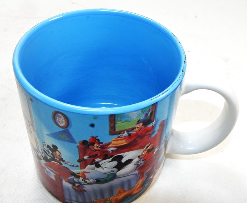 Puede incluir: Una taza de cer&aacute;mica blanca con interior azul y una imagen colorida de Mickey Mouse y sus amigos. La imagen muestra a Mickey Mouse y sus amigos en un dormitorio.