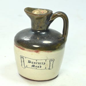 Puede incluir: Una pequeña jarra de cerámica con asa y boquilla. La parte superior y el asa son de color marrón oscuro, que pasa a un color crema más claro en la parte inferior. La jarra tiene una etiqueta que dice "Bunratty Mead".