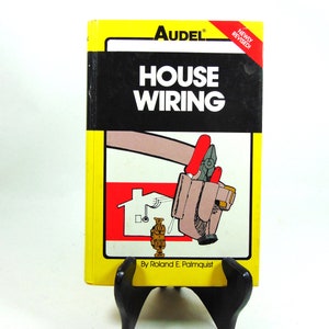 Peut inclure: Un livre jaune intitulé "Audel House Wiring" par Roland E. Palmquist. La couverture présente une illustration de dessin animé d'une maison, une ceinture à outils avec des pinces et une personne tenant un fil.