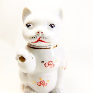 May include: White porcelain cat figurine with red and gold painted details. The cat is sitting with its paw raised, a traditional Japanese lucky cat design.