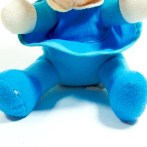 May include: A blue plush toy with a white face and arms. The toy is sitting with its legs spread out.