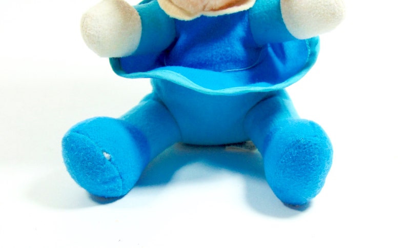 May include: A blue plush toy with a white face and arms. The toy is sitting with its legs spread out.