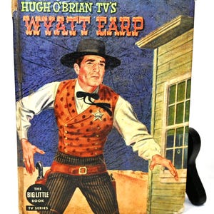 May include: A vintage paperback book cover featuring a color illustration of Wyatt Earp, a lawman from the American Old West, wearing a cowboy hat, vest, and gun belt. The book title is "Hugh O'Brian TV's Wyatt Earp" and the book series is "The Big Little Book TV Series".