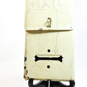 May include: A vintage off-white metal mailbox with the word "MAIL" visible. The mailbox has a hinged door, a slot, and a decorative black stand. The paint is distressed, giving it a rustic appearance.