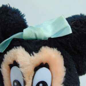 May include: A plush toy of Mickey Mouse with black fur, large ears, and a light blue bow. The toy has large, white eyes with black pupils.