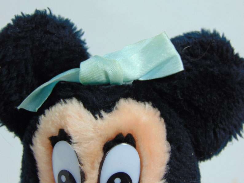 May include: A plush toy of Mickey Mouse with black fur, large ears, and a light blue bow. The toy has large, white eyes with black pupils.