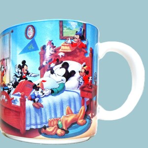 Puede incluir: Una taza de cer&aacute;mica blanca con una ilustraci&oacute;n colorida de Mickey Mouse y sus amigos en un dormitorio. Mickey est&aacute; dormido en la cama, mientras que otros personajes est&aacute;n a su alrededor. La taza tiene un asa en el lado derecho.