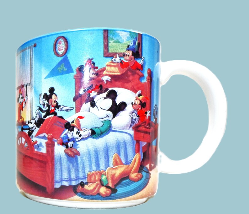 Puede incluir: Una taza de cer&aacute;mica blanca con una ilustraci&oacute;n colorida de Mickey Mouse y sus amigos en un dormitorio. Mickey est&aacute; dormido en la cama, mientras que otros personajes est&aacute;n a su alrededor. La taza tiene un asa en el lado derecho.