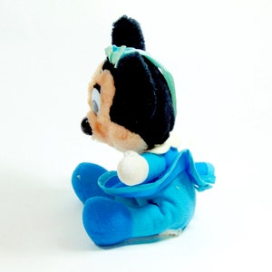 May include: A plush toy of a cartoon mouse wearing a blue dress and a blue bow. The toy is sitting on a white surface.
