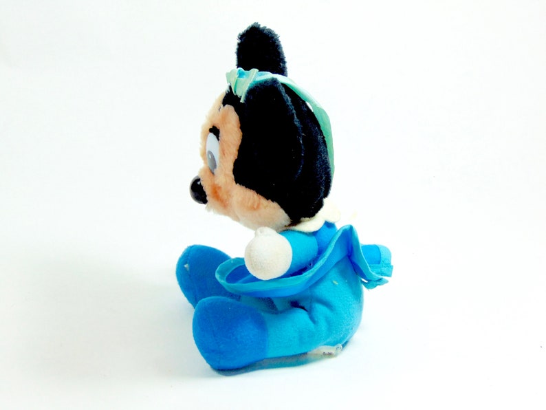 May include: A plush toy of a cartoon mouse wearing a blue dress and a blue bow. The toy is sitting on a white surface.