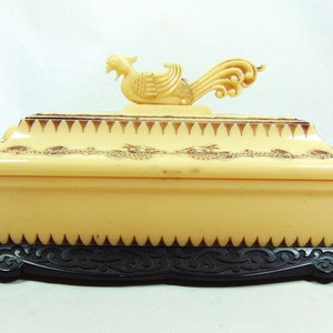 Op de afbeelding: Een crèmekleurige sieradenkistje met een zwarte basis. De kist heeft een decoratieve gouden rand en een deksel in de vorm van een vogel.