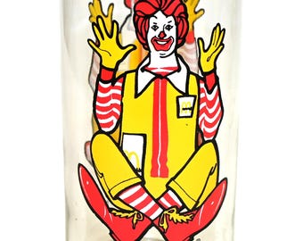 May include: Clear glass featuring a seated Ronald McDonald character in a yellow outfit with red and white striped sleeves and socks. The glass has the text "RONALD MC DONALD COLLECTOR SERIES".