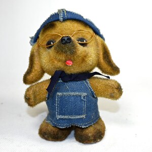 May include: A brown plush toy dog wearing a blue denim hat, overalls, and gold-rimmed glasses. The dog has a red tongue sticking out.