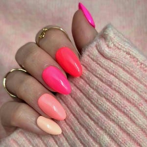 Peut inclure: Gros plan sur une main avec de longs ongles ovales peints dans des tons vifs de rose et de corail. Les ongles sont brillants et réfléchissent la lumière. Des bagues dorées ornent les doigts. La main est partiellement couverte par un pull en tricot rose clair.