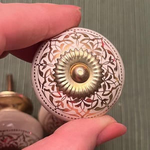 May include: White and gold decorative cabinet knobs with intricate floral patterns. The knobs have a round shape with a gold center and a brass stem. The knobs are likely made of ceramic or porcelain.