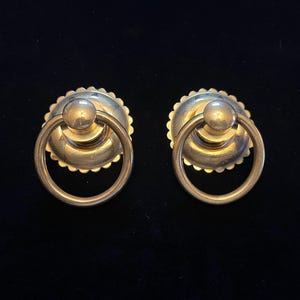 May include: Two antique brass door pulls. Each pull features a circular backplate with a scalloped edge and a central ring handle. The brass has a warm, aged patina, suggesting vintage craftsmanship. The pulls are set against a dark background.