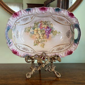 May include: An oval, antique porcelain serving dish with a hand-painted grape design. The dish has a scalloped edge with pink, blue, and white accents, and gold trim. It is displayed on a decorative brass stand.