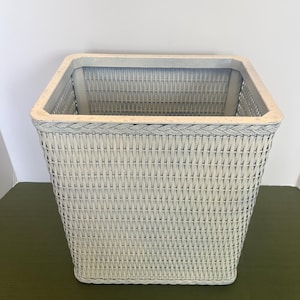 May include: A square, off-white wicker basket with a woven design. The basket has a light gray trim around the top and a braided border. The basket is sitting on a green surface.