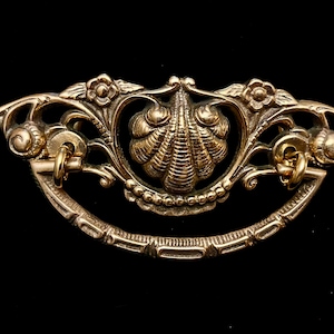 May include: Ornate gold-toned drawer pull with a seashell design at the center. The pull features floral and scrollwork details, with a curved handle and small rings for attachment. The hardware is set against a black background.