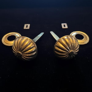 May include: Two antique brass cabinet knobs with a ribbed design, each with a matching backplate and square mounting hardware. The knobs are set against a dark background.