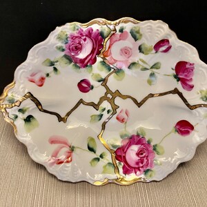 May include: A white porcelain serving dish with a scalloped edge and gold trim. The dish is decorated with pink and red roses and green leaves. The dish is cracked in several places, with the cracks filled in with gold paint.