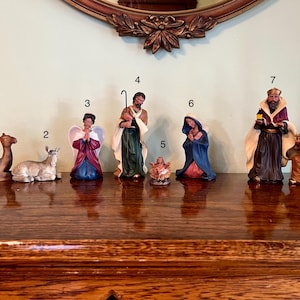 May include: A collection of ceramic nativity figurines, including a camel, donkey, angel, Joseph, baby Jesus, Mary, and the three wise men. The figures are painted in various colors, with detailed clothing and facial features. The set is displayed on a wooden surface.